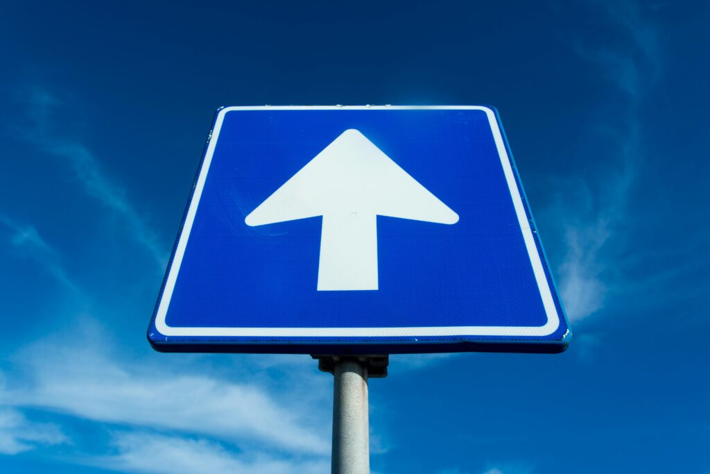 Blue traffic sign with an upward white arrow pointing up, set against a clear blue sky.